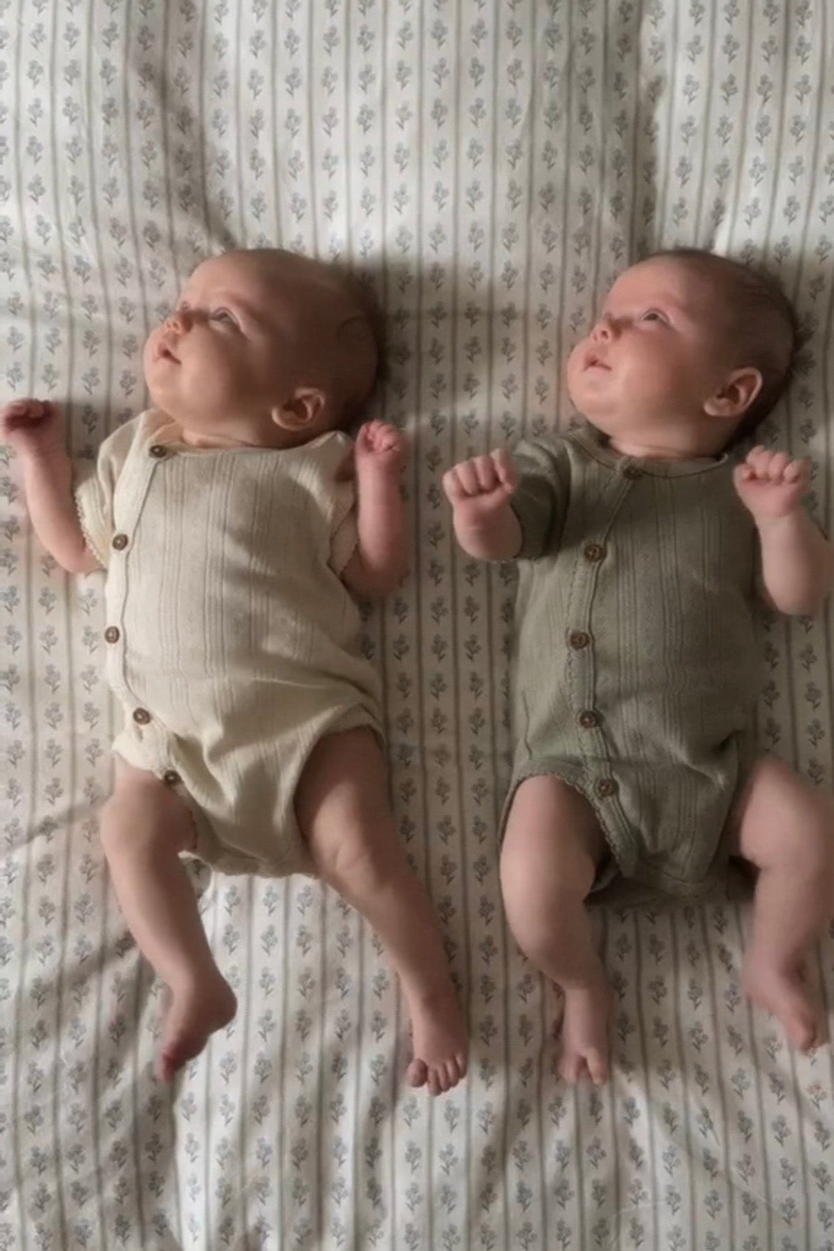 Two babies in green and beige vests lying on a patterned duvet cover.